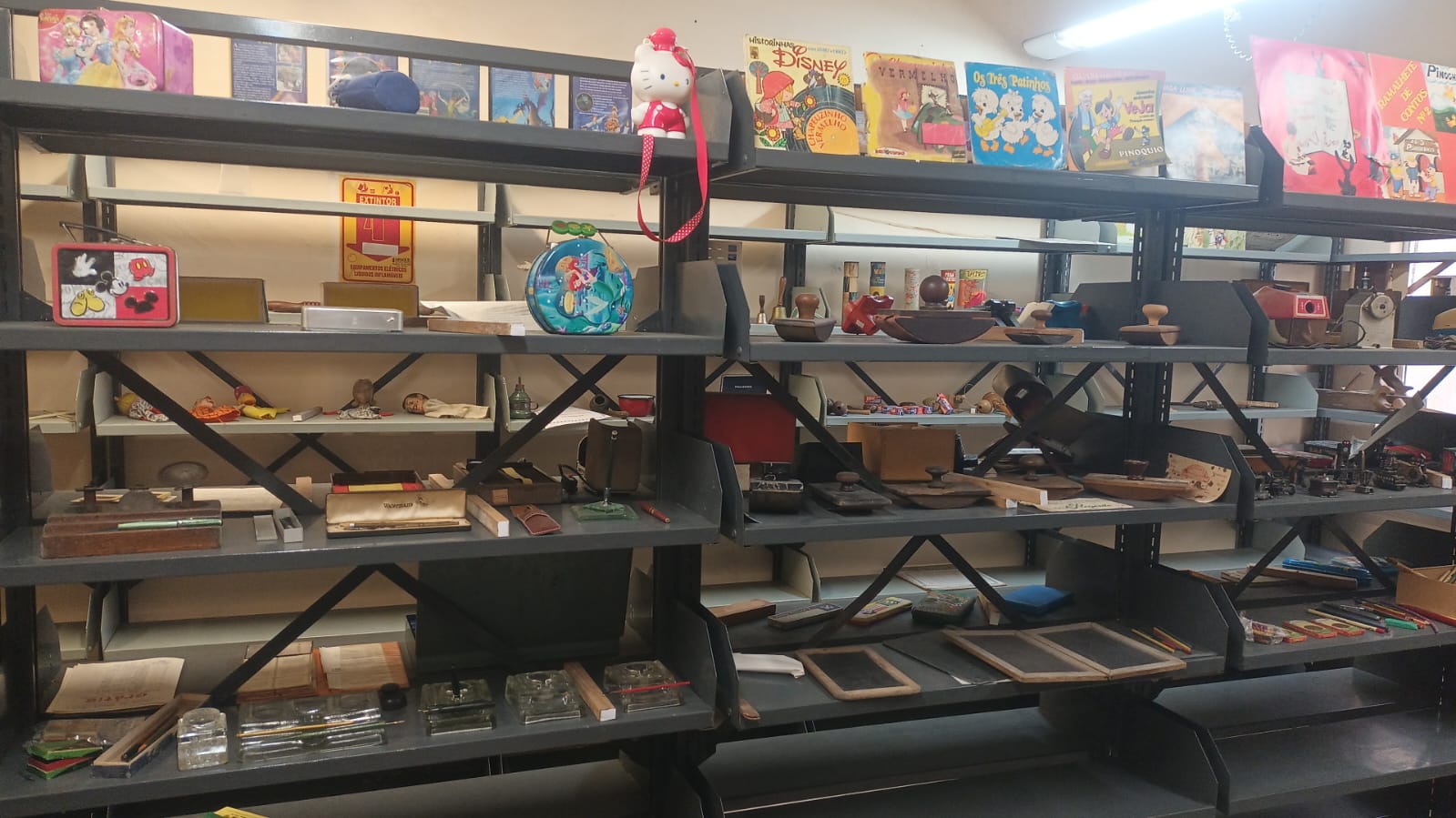 Exposição Cedoc com uma estante de metal cinza com quatro prateleiras cheias de objetos antigos e livros.