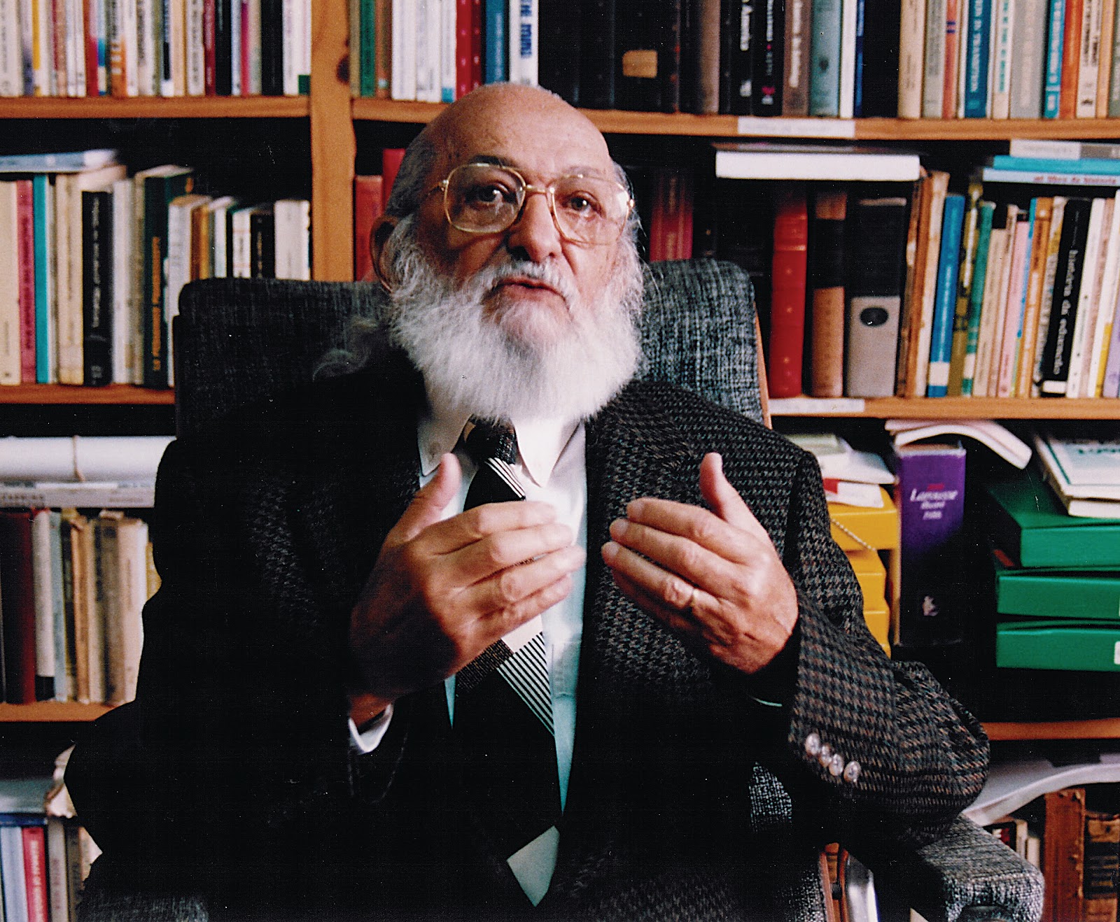 Imagem de Paulo Freire sentado, ao fundo encontra-se uma estante de madeira com diversos livros. Paulo Freire é um homem idoso, careca, de barba grande branca, usa óculos e terno preto listrado com preto; usa gravata preta e camisa social branca.
