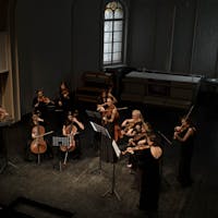 A diverse group of women performs classical music with violins and cellos on an indoor stage.