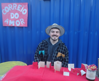 Indivíduo branco em traje típico de festa junina em um stand com cartões postais, sob um cartaz Correio do Amor.