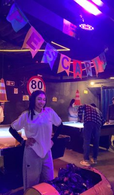 Uma jovem, de pele morena clara e cabelos escuros e lisos até os ombros, sorri para a câmera. Ela veste uma camiseta polo branca de manga curta sobre uma blusa preta de manga longa, calça jeans clara rasgada nos joelhos e está com as mãos na cintura. Ela se posiciona na frente de uma mesa de sinuca esverdeada e um tanto desgastada, que ocupa grande parte da parte inferior esquerda da imagem. À sua direita e ao fundo, um homem de camisa xadrez está curvado sobre outra mesa de sinuca jogando, e um segundo homem de casaco escuro está de pé mais ao fundo. À esquerda, outro homem de blusa branca está de pé ao lado de outra mesa de sinuca. O ambiente é escuro, iluminado por luzes neon em tons de roxo e rosa que refletem no teto e nas paredes. Acima das mesas, bandeirinhas coloridas com letras que formam a palavra "PESCARIA" estão penduradas. Há cones de trânsito laranja e brancos e placas de sinalização (uma com "80" e outra com setas indicando saída) espalhados pelo local. Barris de metal enferrujados também são visíveis, um deles na parte inferior direita com o que parecem ser sacos de lixo pretos.