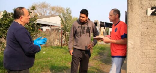 Professor Alejandro está mais à esquerda da imagem com blusão azul, calça preta e luvas de plástico azul. Ele está conversando com dois homens que estão mais à direita, um com moletom cinza e calça preta, e outro com camiseta vermelha de detalhes azuis e shorts cinza segurando uma prancheta. Os três conversam próximos à casa com número 21.