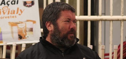 No centro da imagem, Alexandre Quadrado: um homem na faixa dos 50 anos, com cabelos escuros e barba fechada. Ele veste um casaco preto de abrigo, e está de perfil — como quem conversa com outra pessoa.Nos fundos, grades brancas com um cartaz publicitário pendurado.