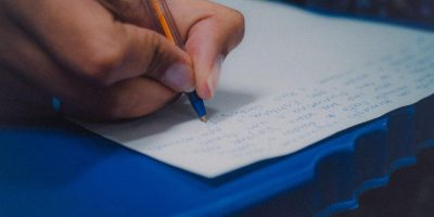 A foto mostra em plano detalhe a mão de uma pessoa escrevendo em um papel, sobre uma mesa azul.