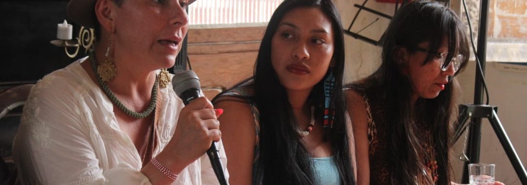 A foto mostra três mulheres indígenas sentadas lado a lado, participando de uma conversa ou roda de diálogo em um ambiente interno iluminado pela luz natural que entra por uma janela. À esquerda, está uma mulher de pele clara e cabelo curto, usando um chapéu cinza com pena colorida, brincos grandes dourados e um colar verde. Ela veste uma blusa branca de mangas largas e segura um microfone com uma de suas mãos enquanto fala. Sua expressão é séria e focada. No centro, há uma mulher jovem de cabelos longos e lisos, pele morena e expressão tranquila. Ela observa atentamente a mulher que fala. Usa um vestido azul claro e longos brincos coloridos e artesanais pendurados. Suas mãos repousam no colo. À direita, está uma mulher de cabelos longos e escuros, usando óculos, um vestido com estampa animal print, brincos longos e colares. Ela segura um copo de água nas mãos e olha para baixo, como se estivesse refletindo. Ao fundo, aparecem elementos do espaço: paredes antigas, madeira, um extintor vermelho e parte de uma estrutura metálica. O clima da cena transmite escuta, troca e presença coletiva.