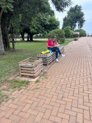 Mulher adulta sentada em um banco modular de madeira em uma área externa arborizada, semelhante a um parque ou praça. Ela veste camiseta rosa, calça jeans azul e tênis claros. Está com a cabeça inclinada para baixo, concentrada em um caderno ou prancheta apoiado no colo, preenchendo planilhas. Ao lado dela, sobre o banco, há uma bolsa amarela. O entorno é composto por gramado, árvores de grande porte ao fundo e um caminho pavimentado com blocos intertravados à direita.