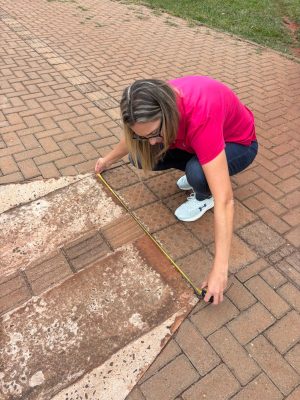 Mulher adulta agachada sobre uma calçada de blocos intertravados, realizando a medição do piso com uma trena amarela. Ela veste camiseta rosa, calça jeans azul, tênis claros e óculos. A trena está estendida sobre uma faixa de piso diferente, formada por placas retangulares antigas e desgastadas, com textura irregular e manchas, contrastando com o pavimento ao redor. A mulher segura a trena com atenção, observando a medida, e ao fundo é possível ver área gramada adjacente à calçada.