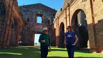 Homem e mulher adultos caminham por entre ruínas de uma antiga edificação de pedra, com grandes arcos e paredes altas parcialmente preservadas. O homem veste chapéu claro, óculos escuros, blusa verde e calça escura, e faz um gesto com a mão enquanto conversa. A mulher veste camiseta azul, calça jeans e óculos escuros, caminhando ao lado dele com as mãos nos bolsos. O chão é coberto por gramado verde e, ao fundo, outras pessoas aparecem pequenas, caminhando pelo espaço. O céu está azul e a iluminação indica um dia ensolarado.