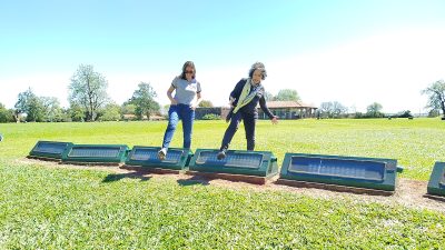 Duas mulheres adultas caminham sobre uma sequência de equipamentos baixos e retangulares instalados no gramado, alinhados em fila. Os equipamentos são verdes, com superfícies envidraçadas ou painéis na parte superior. A mulher à esquerda veste camiseta clara e calça jeans, observando onde pisa. A mulher à direita veste roupa escura e um lenço claro no pescoço, abrindo os braços levemente. Ao fundo, há um amplo campo gramado, árvores espalhadas e edificações baixas, sob um céu claro e ensolarado.