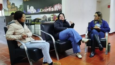 Três mulheres adultas sentadas em poltronas pretas, dispostas em semicírculo, em um ambiente interno com piso cerâmico avermelhado. Elas participam de uma conversa ou reunião. A mulher à esquerda veste jaqueta clara e calça jeans, segura um caderno no colo e olha em direção às outras. A mulher ao centro veste jaqueta escura e calça azul, está sentada com as pernas cruzadas e gesticula com uma das mãos enquanto fala. A mulher à direita veste moletom azul, calça escura e botas pretas, segura uma prancheta e observa atentamente. Ao fundo, há painéis e fotografias expostas na parede, incluindo imagens de edificações históricas e paisagens.